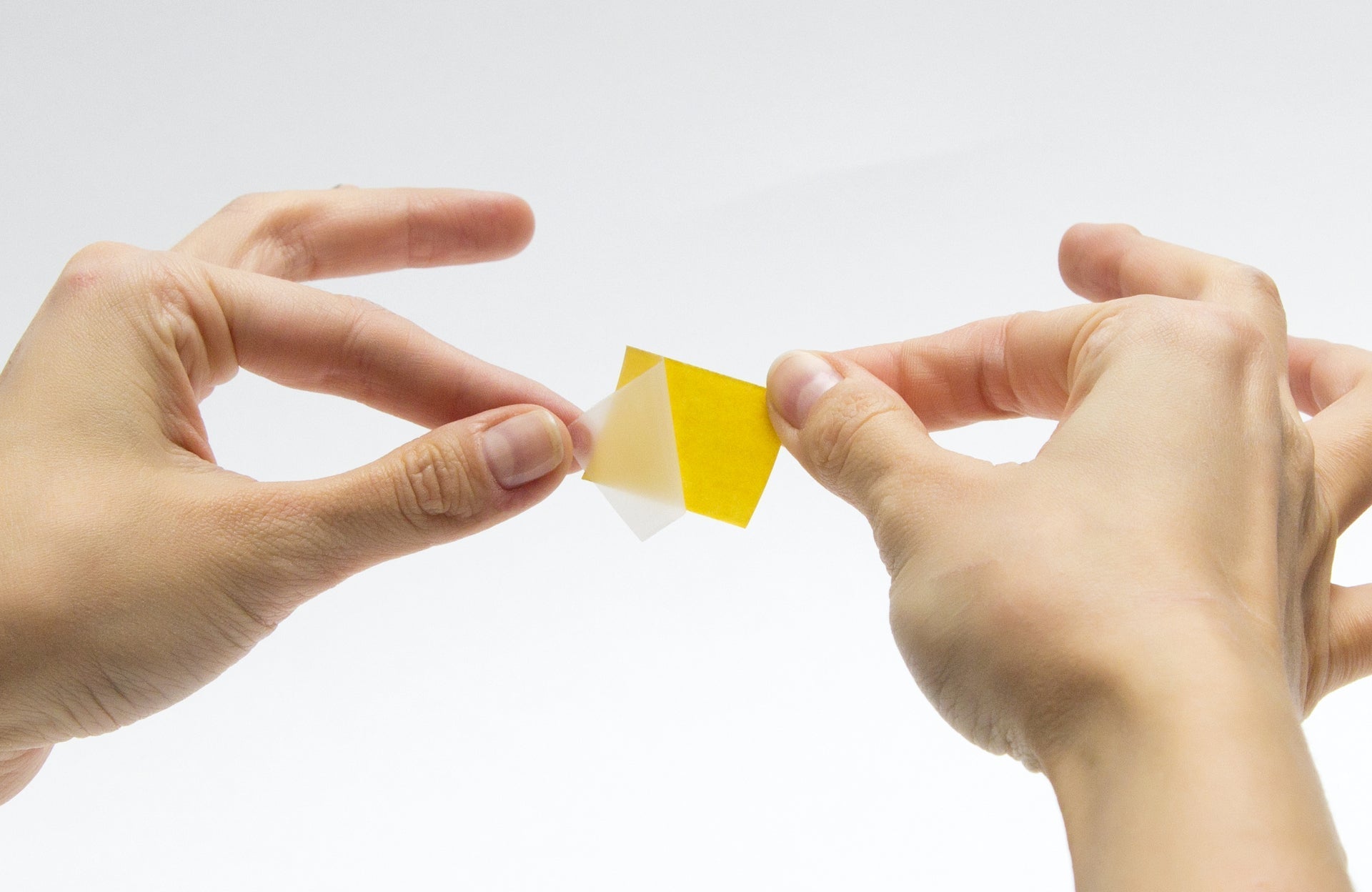 A close-up of a person peeling the yellow backing paper off a fashion tape strip, holding the strip firmly with their other hand